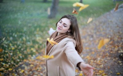 Femme en beige, bras tendus, yeux fermés. Feuilles d'automne, nature floue. Bonheur paisible, journée lumineuse.